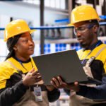 In smart manufacturing plant, african american engineers supervise industrial automation system using laptop. The automated factory features conveyor belts, robotic arms and optimized assembly lines.
